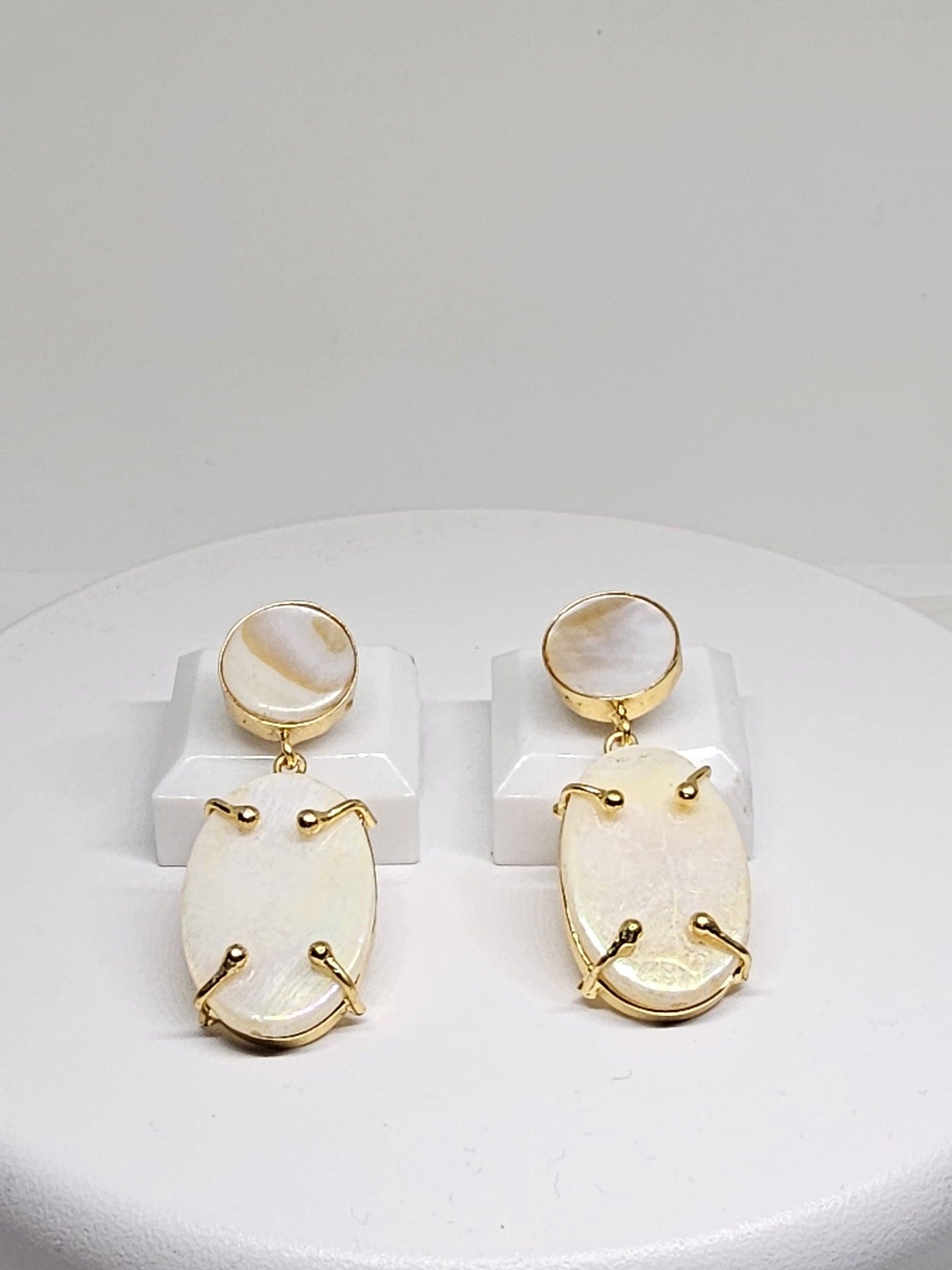 Gold-plated dangle earrings with oval-shaped Mother of Pearl stones on a white display stand