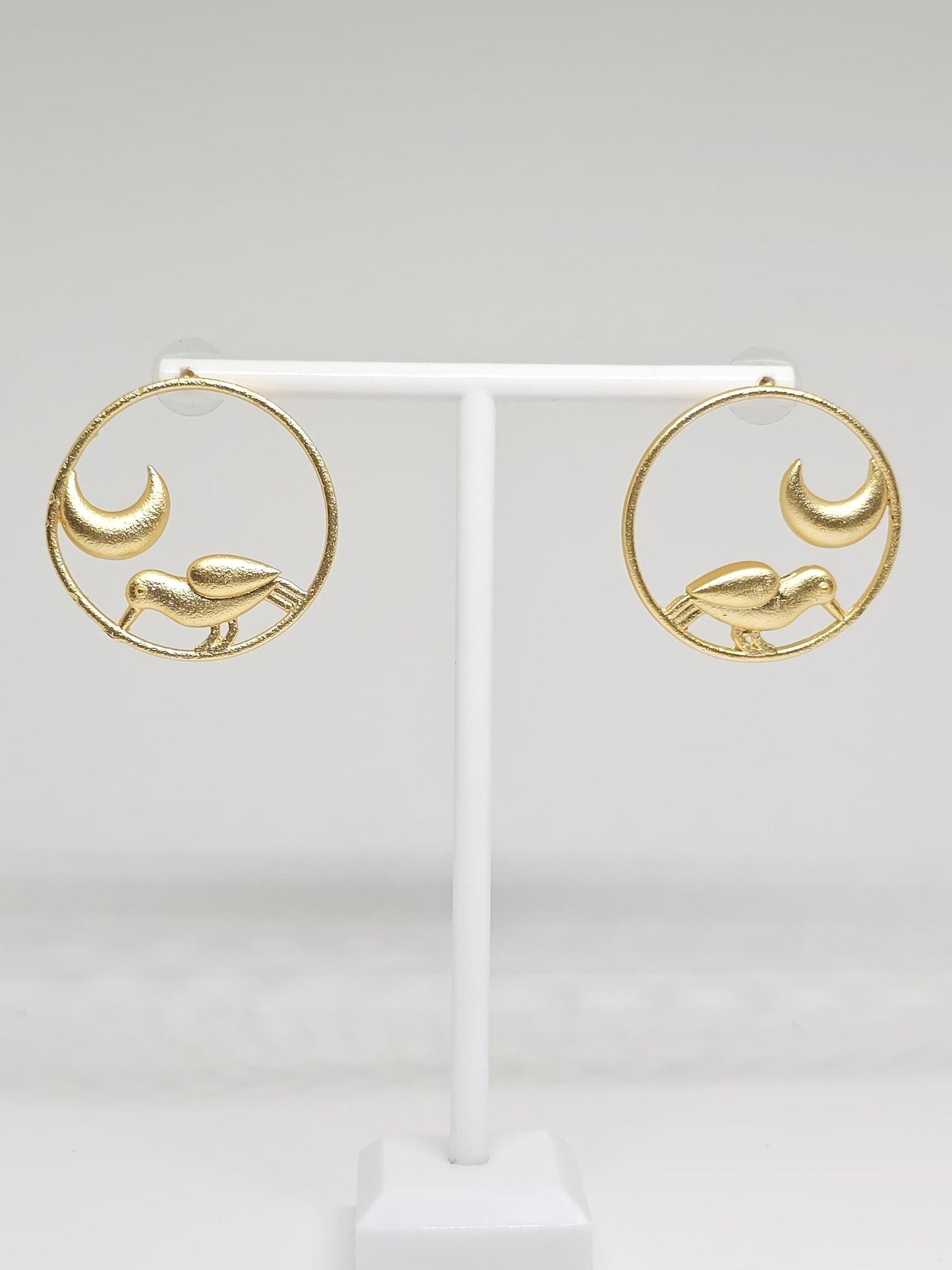 Brass hoop earrings with bird and crescent moon design on display stand.