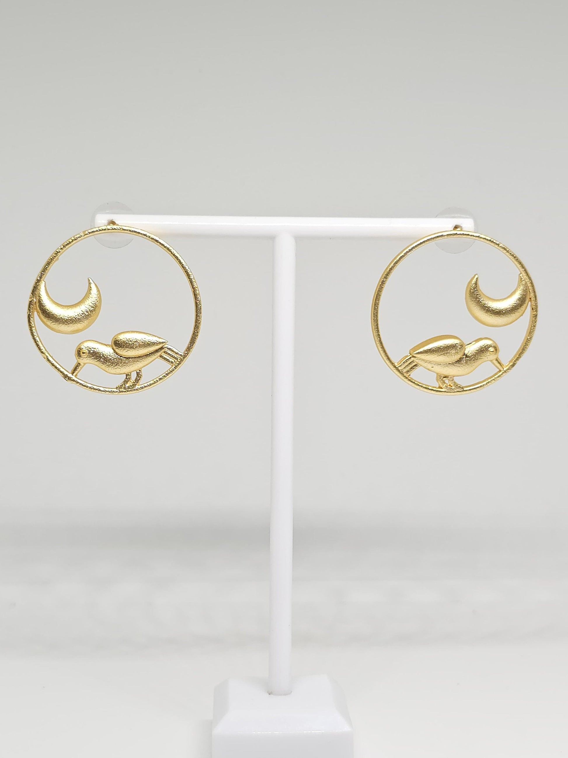 Brass hoop earrings with bird and crescent moon design on display stand.